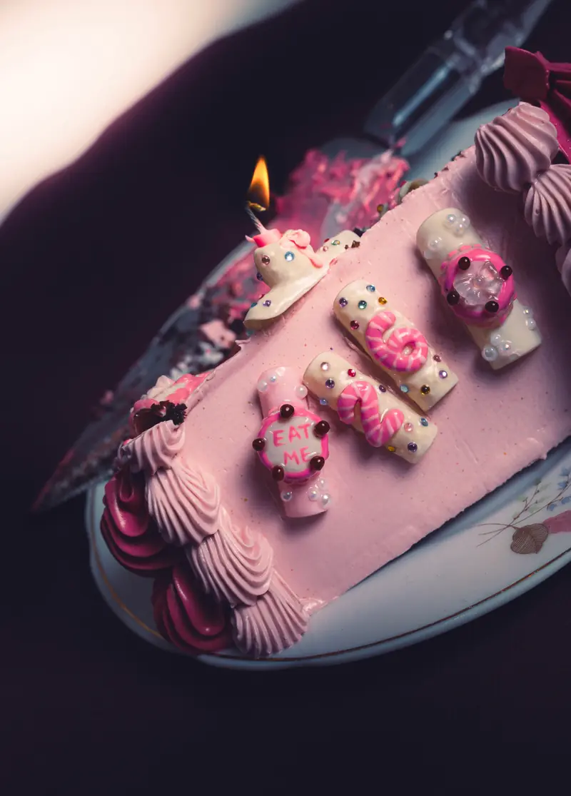 Bolo rosa com unhas decoradas em 3D sobre a cobertura, formando o número 26, e uma vela acesa
