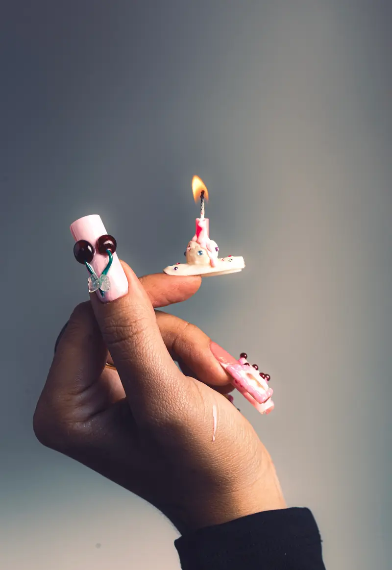 Mão segurando uma unha rosa com cerejas e laço de vidro, com outra unha em formato de bolinho com vela acesa na ponta do dedo