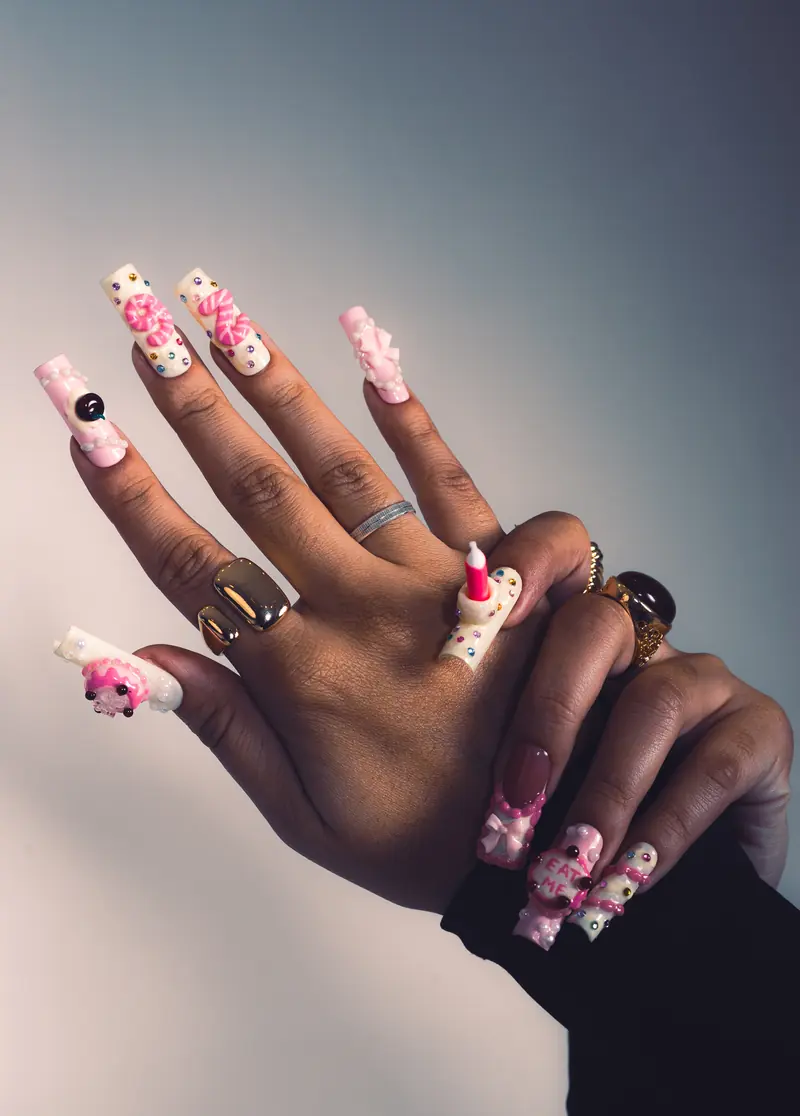Mãos da Beatriz Concept com unhas longas decoradas em 3D: números em relevo, laço, cerejas e uma vela de aniversário sobre a unha